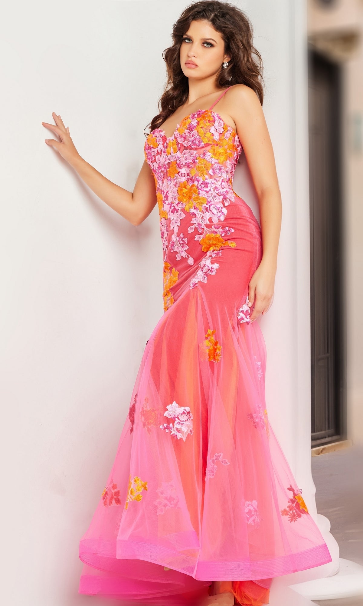 A woman stands against a white wall, looking slightly away, wearing the Long Prom Dress 36843 by Jovani, a pink and orange mermaid tulle gown with intricate floral embroidery.